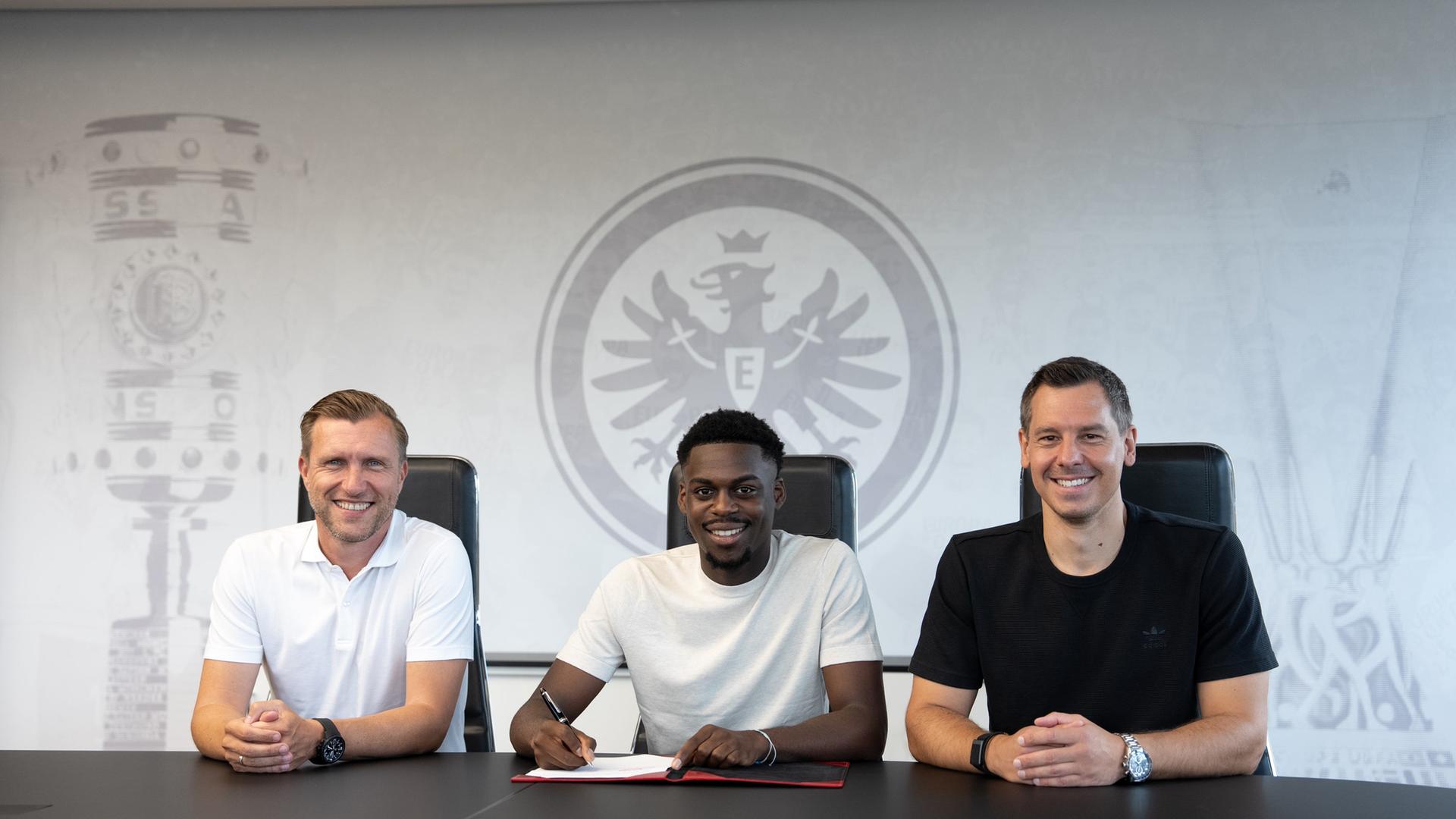 Noel Aseko unterschreibt, umgeben von Markus Krösche und Timmo Hardung.  | Eintracht